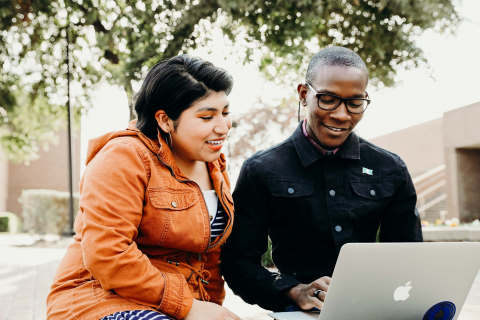 Two individuals looking at a silver Apple Laptop. One is a woman in orange and the other is a male in a dark navy jacket.