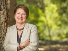 Joanna Regulska's headshot. Joanna is leaning against a tree smiling with her arms crossed in front of a leafy green background. She is wearing a tan jacket.