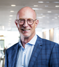 Graham Carr's headshot. Graham is standing in front of a blurred out office room, wearing a blue plaid suit jacket with a light blue shirt.