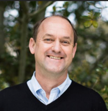 David Ackerly's headshot. David is standing in front of leafy green trees and wearing  a dark blue sweater.