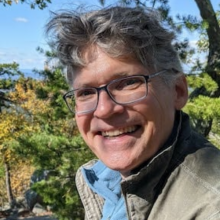 Bruce has gray hair and glasses, and is standing in front of a blue sky and green trees, he is wearing a green jacket with a blue shirt