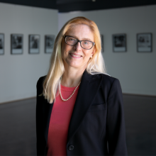 Cinzia has blonde hair and is standing in an art gallery wearing a black blazer and a pink shirt, she is smiling