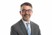 Eric Leshinskie's headshot. Eric is in front of a white background wearing a gray suit jacket  and a blue tie. 