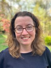 Leah Nichols' headshot. Leah is wearing glasses and standing in front of leafy green trees, she is wearing a dark blue sweater