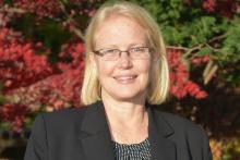 Joanie's headshot- Joanie is wearing glasses, a black suit jacket, and is standing in front of a japanese maple tree with red leaves