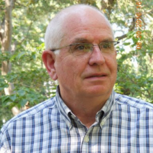 Richard has gray hair and glasses and a blue plaid shirt. He is standing in front of some green trees.