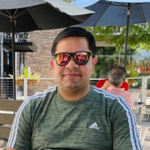 Rajesh is sitting outside at a caffe, wearing sunglasses and a green shirt