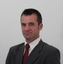 Stephane's headshot. He is in front of a white background and wearing a dark gray suit with a red tie. 